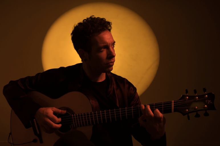 Pipo Romero plays the guitar while looking to the side with a yellow circle background